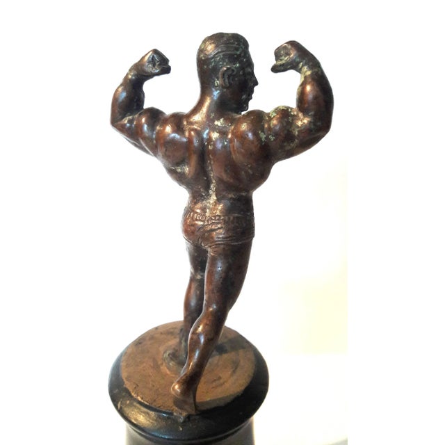 A small bronze muscle man statue on a black wooden base. Measures: 9 1/2" high x 4" diameter base.