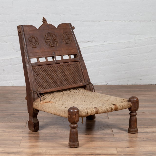 An old Indian rustic wooden low chair from the early 20th century, with hand-woven twine and carved motifs. Charming our...