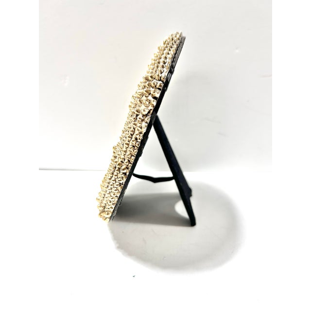 A sculptural round picture frame featuring a dramatic field of raised metallic studs in a warm gold finish. The heavily...
