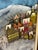 This artwork is a mixed-media/3D collage painting depicting a whimsical winter village built into a snowy, mountainous...