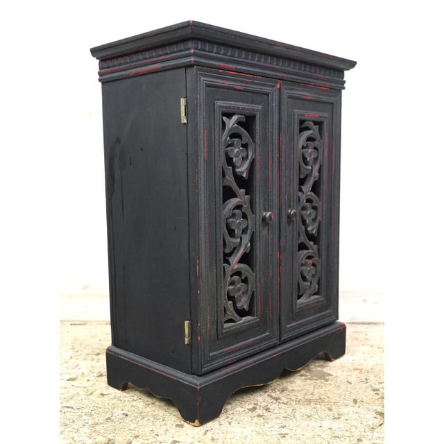 Vintage tabletop storage cabinet made of black painted wood with red distressed highlights. It features two carved cutout...