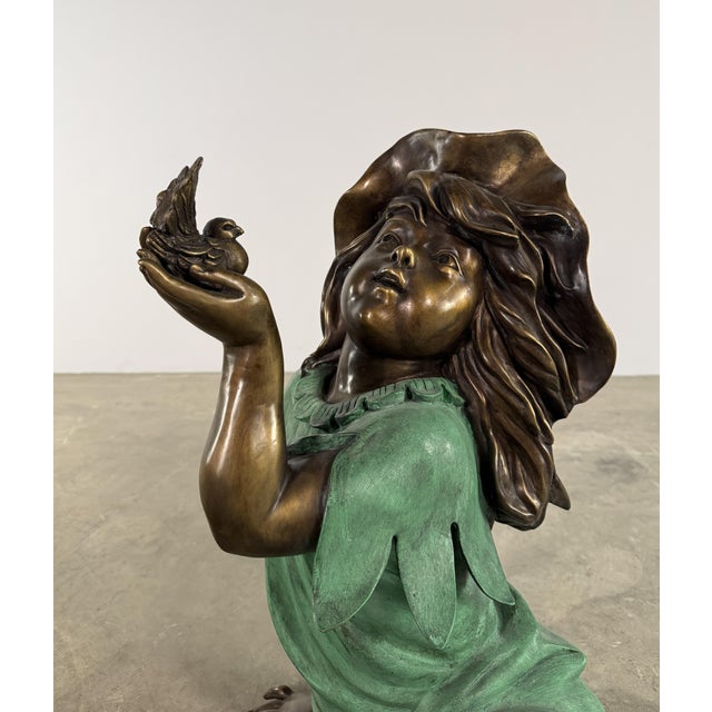 This enchanting bronze sculpture depicts a young girl seated in a contemplative pose, gently holding a small bird in her...