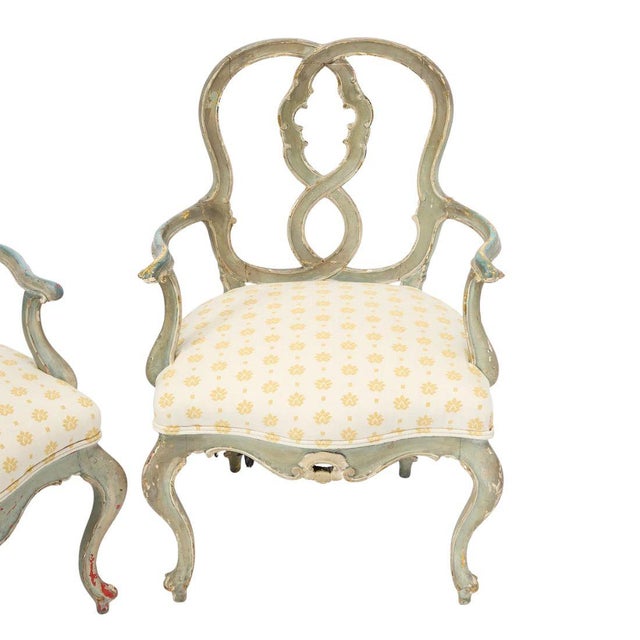 19th century pair of armchairs featuring a gracefully carved wooden frame with a softly distressed, pale green painted...