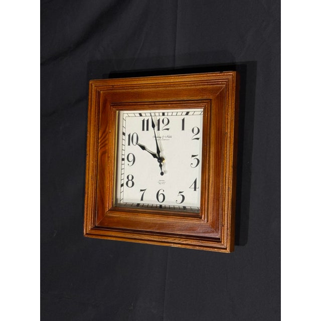 A handsome square wall clock by Sterling & Noble Clock Company, featuring a warm wood frame, classic ivory dial, bold...