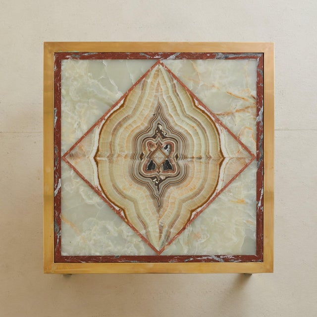 An onyx coffee table dating to the 1970s, its square brass frame enclosing an inlaid onyx top. At the center, four...