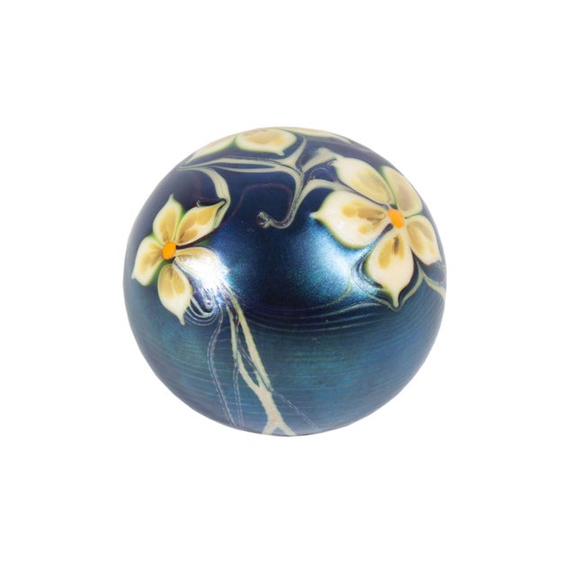 A 1976 iridescent art glass paperweight by Orient and Flume. This spherical paperweight features yellow flowers against a...