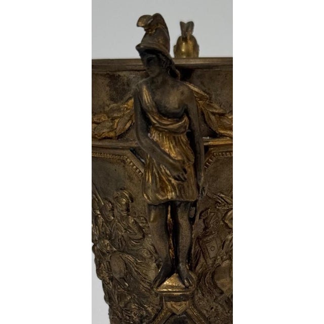 A richly detailed Neoclassical bronze cup-form sculpture featuring dramatic high-relief battle scenes and three fully...