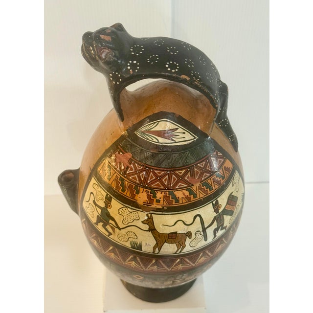 A dramatic hand-painted South American folk art pottery vessel featuring a sculptural jaguar handle gracefully arching...