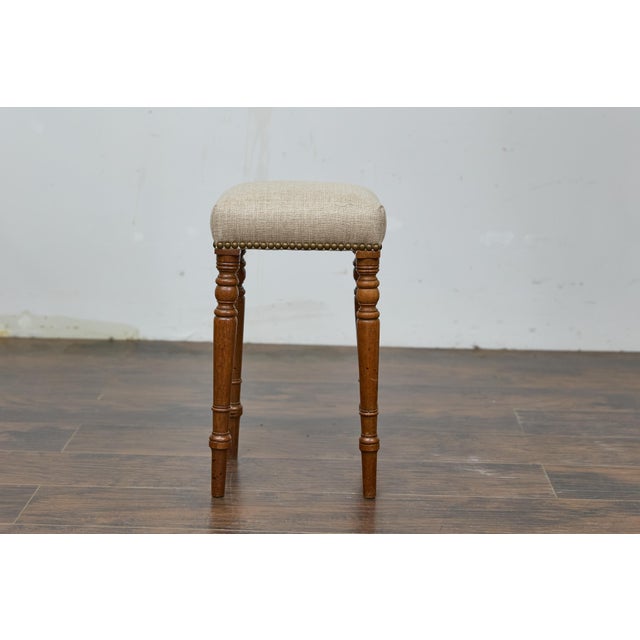 An English petite oak stool from the 19th century featuring custom linen upholstery and turned legs. The stool is...