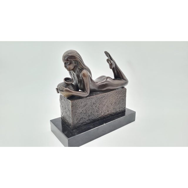 A bronze sculpture depicting a naked woman lying down and holding a vase or jug, identified as the figure of Aquarius. The...