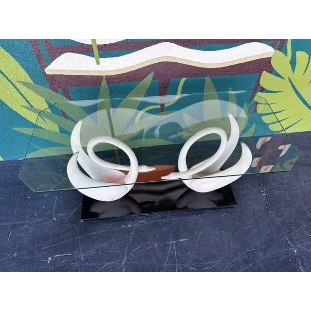 Console table has a glass top which rests atop two composite resin ivory colored swans that sit on a wood and black...