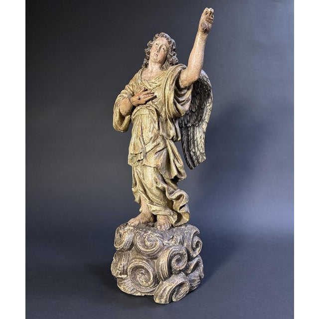 The polychrome wood carved sculpture depicts an angel standing on a pedestal of clouds, his left hand on his chest and his...