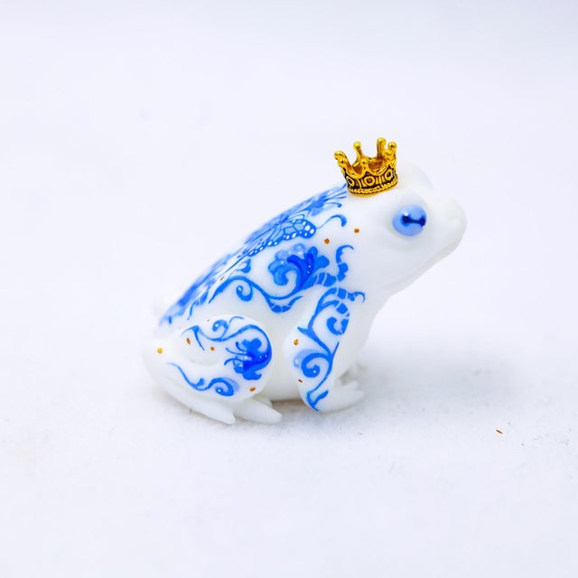 This artwork features a small, intricately designed frog, adorned with blue and white patterns reminiscent of Delft...