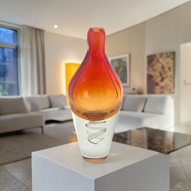 A sculptural sommerso art glass vase featuring a rich amber-orange upper casing suspended over a clear base, accented by...