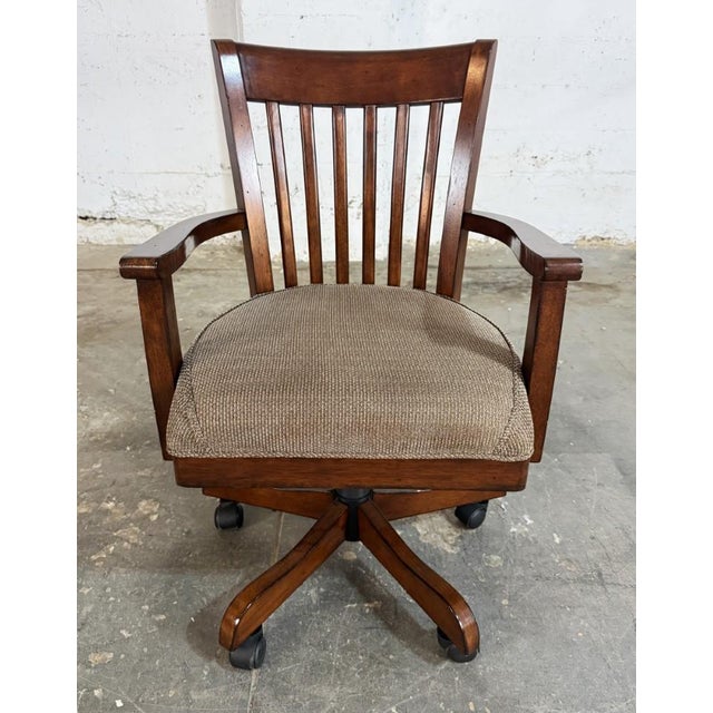 This traditional desk chair by Riverside Furniture features a warm wood frame, vertical slat back, curved arms, and a...