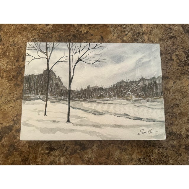 This original watercolor captures the stillness of a winter landscape, with bare trees standing against softly layered...