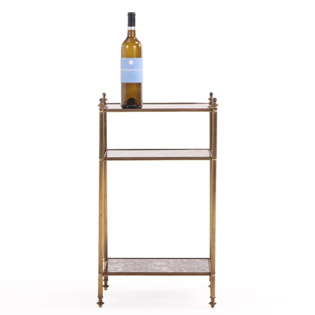 The table features a refined brass frame with neoclassical detailing and three tiers of inset mottled glass shelves,...