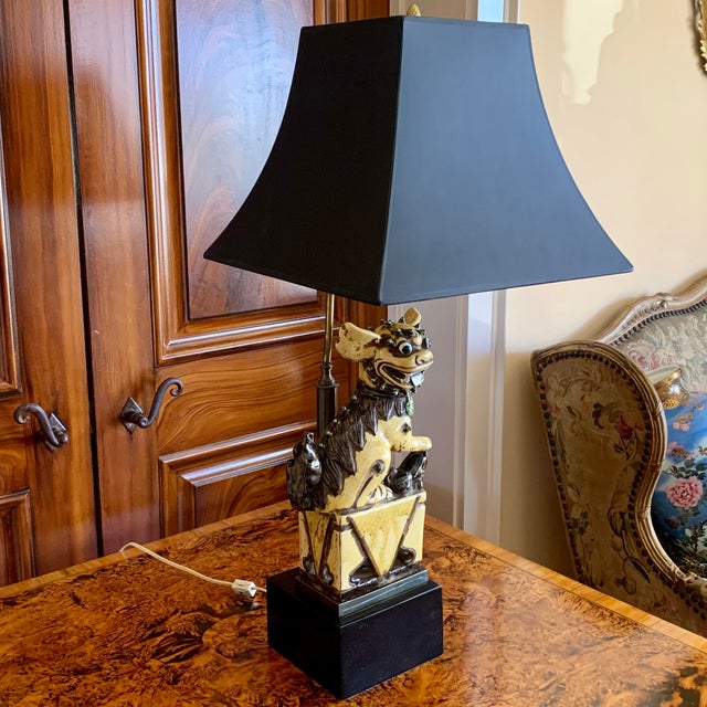 A whimsical grinning yellow glazed ceramic foo dog, with big ears and a spiny back, resting on a plinth, and holding a paw...