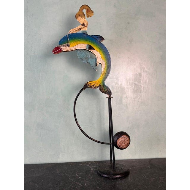 A whimsical vintage folk art kinetic sculpture depicting a young rider perched atop a gracefully arched dolphin. Crafted...