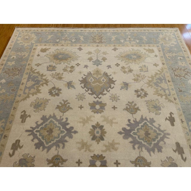 This 9'3" x 11'10" (111" x 142") Indian Oushak rug features a calming palette of soft blue, ivory, taupe, and sand, woven...