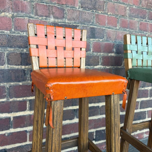 This pair of Ranch Oak bar stools have orange and green genuine leather seats with wrapped tasseled corners and woven seat...