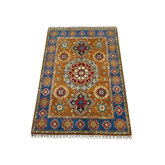 A warm rust field centers a bold Kazak medallion, surrounded by smaller motifs in blue, burgundy, cream, sage green, and...