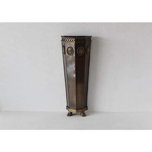 A Mid-century brass and other metal umbrella stand that features an elegant half-hexagonal shape with intricate decorative...