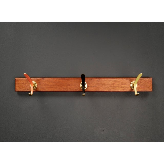 Oak robe rack with beveled edges for wall mounting, featuring two long, rounded brass rods in red, black, and gold. A...