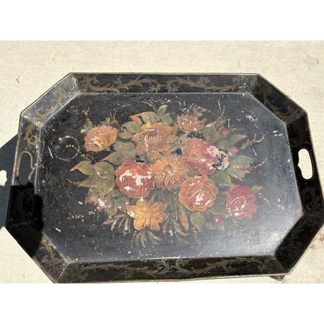 The hand-painted toleware features a colorful, worn floral bouquet in the center of a black metal tray with cutout handles...
