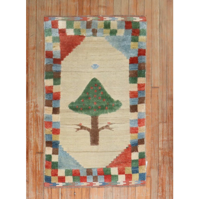 A late-20th century Gabbeh rug with a checkered colorful border surrounded by a stand-alone tree on a camel ground woven...