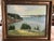A large landscape painting of a lake and the houses and trees surrounding the lake. The painting is signed in the bottom...