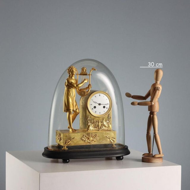 Gilded bronze table clock with enameled metal dial and Roman numerals, flanked by a sculpture depicting a young lyre...