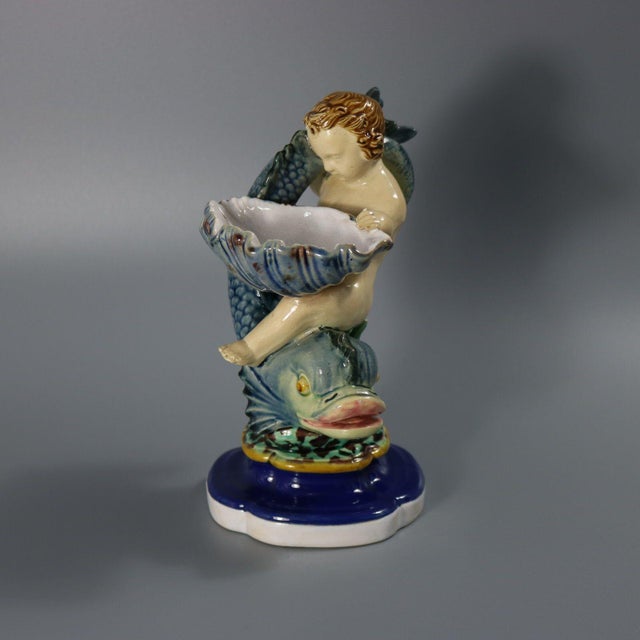 Minton Majolica salt which features a boy holding a shell, climbing over a dolphin. Colouration: grey, cobalt blue, blue,...