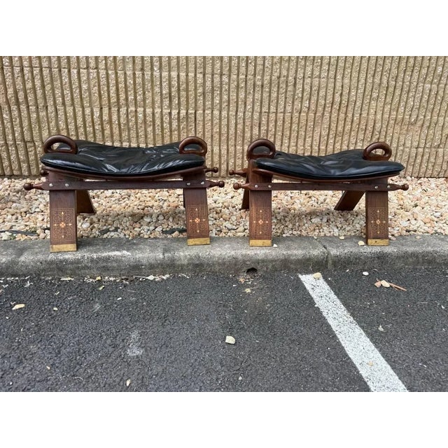 Pair of Vintage Camel Saddle Stools 20th Century, with black vinyl upholstered seat cushion, the base with brass inlaid in...
