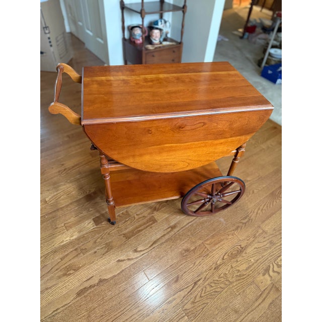 A wooden drop-leaf tea cart featuring an oval top with two folding side leaves, turned spindle legs, a lower shelf, and...