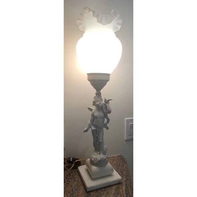Sitting on a marble base, this is a Neoclassical Biscuit Porcelain Putto / Cherub Lamp with Milk Glass Ruffled-edged...