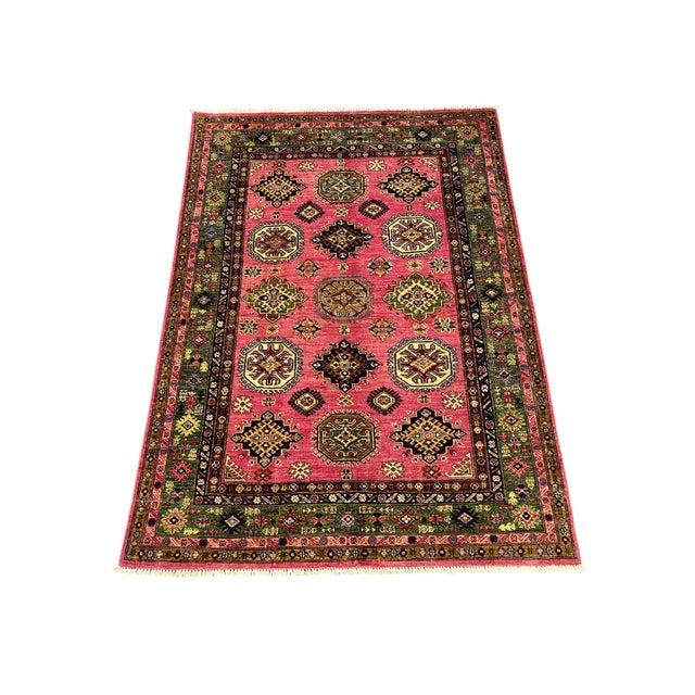 This Kazak-style tribal rug pairs a bright pink field with a deep green patterned border. Repeating motifs in ivory, navy,...