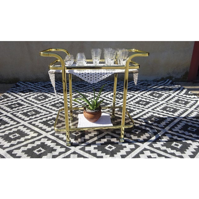 Gold-tone metal serving cart with a flattened tubular frame and smoked glass shelves. Two tempered glass shelves framed by...