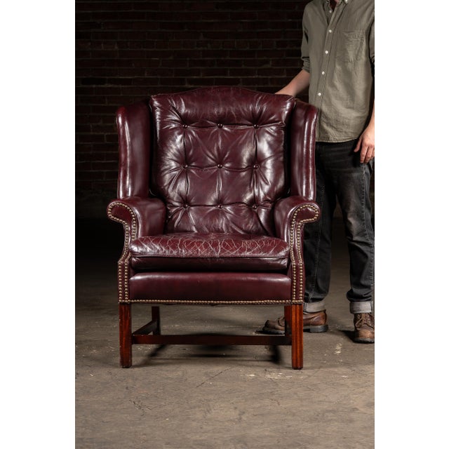 High-back wing chair and matching ottoman upholstered in deep oxblood red leather with brass nailhead trim and mahogany-...