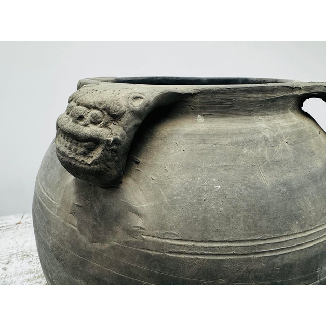 A dark, burnished storage jar featuring two expressive guardian-head handles at the shoulder. The vessel’s broad form and...