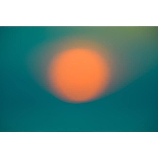 This photographic work features a softly blurred orb of orange, evoking the warmth and fleeting nature of sunset against a...