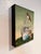 This artwork depicts a weathered fishing boat, partially submerged in lush green grass, with a vibrant hummingbird perched...