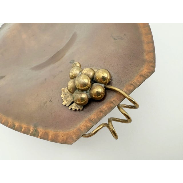 Mixed metal 1970s copper pedestal bowl with brass handles shaped as grapes on vines. The bowl is shallow and sits on top...