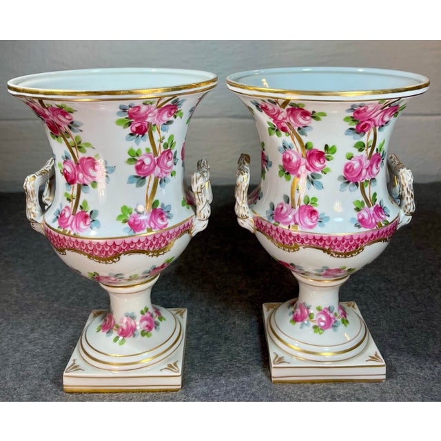 Antique pair of Dresden porcelain urns featuring enameled pink rose depictions alongside gilt accents, double loop...