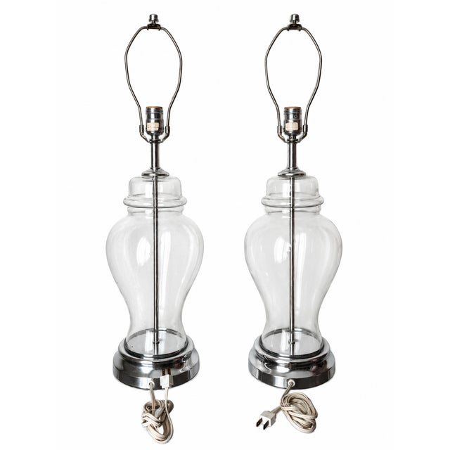 An elegant matched pair of table lamps featuring clear glass bodies with rounded shoulders, gently cinched waists, and...