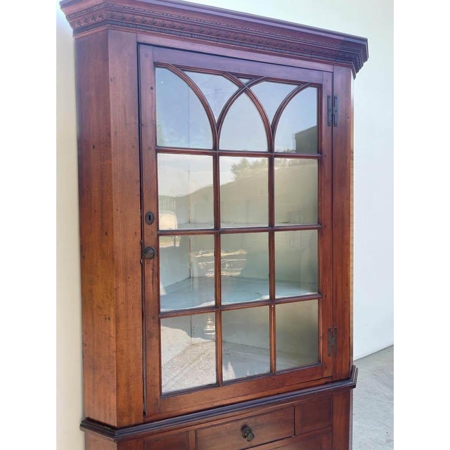 Beautiful antique wood corner cabinet featuring a wavy glass-paneled upper display with Gothic arch mullions, decorative...