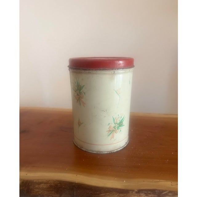 Charming vintage tin canister featuring a delicate floral motif on a soft cream background, topped with a classic red lid....
