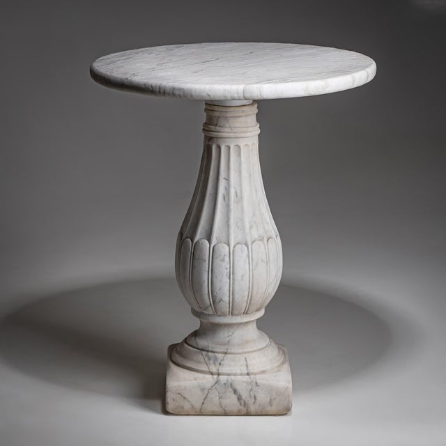 Classical table in white marble featuring a baluster-shaped column and a round top. The surface shows traces of natural...