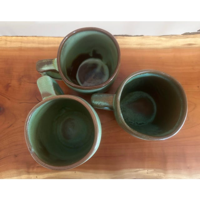 A charming set of three vintage Frankoma stoneware mugs featuring a rich green drip glaze finish with earthy brown...