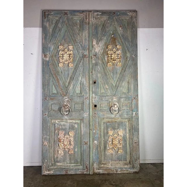 Pair of tall, rustic double doors with an ornate and distressed finish. The doors are painted in a faded greenish-blue...